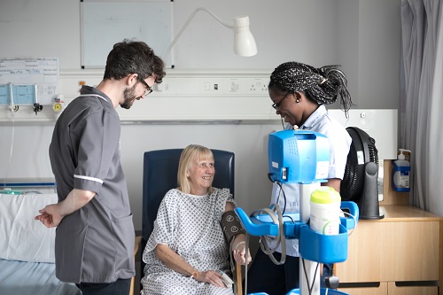 A patient in a hospital gown smiles while interacting with two healthcare professionals